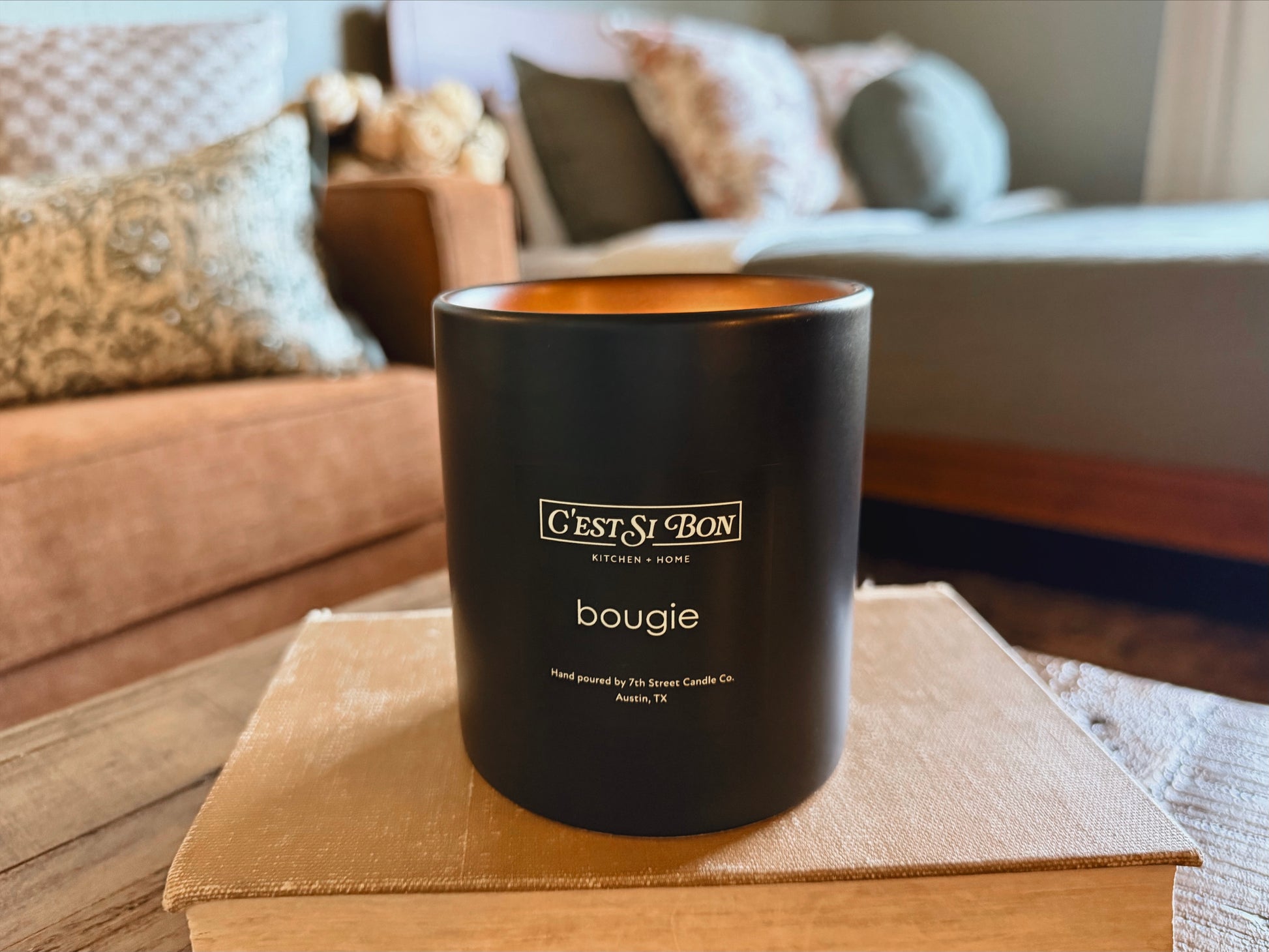 C'est Si Bon-branded black candle in the scent Bougie lit on a wooden surface with a blurred hotel room background.