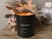 Black candle labeled 'Cést Si Bon' in the scent 'Heather' on a wooden surface with decorative elements.