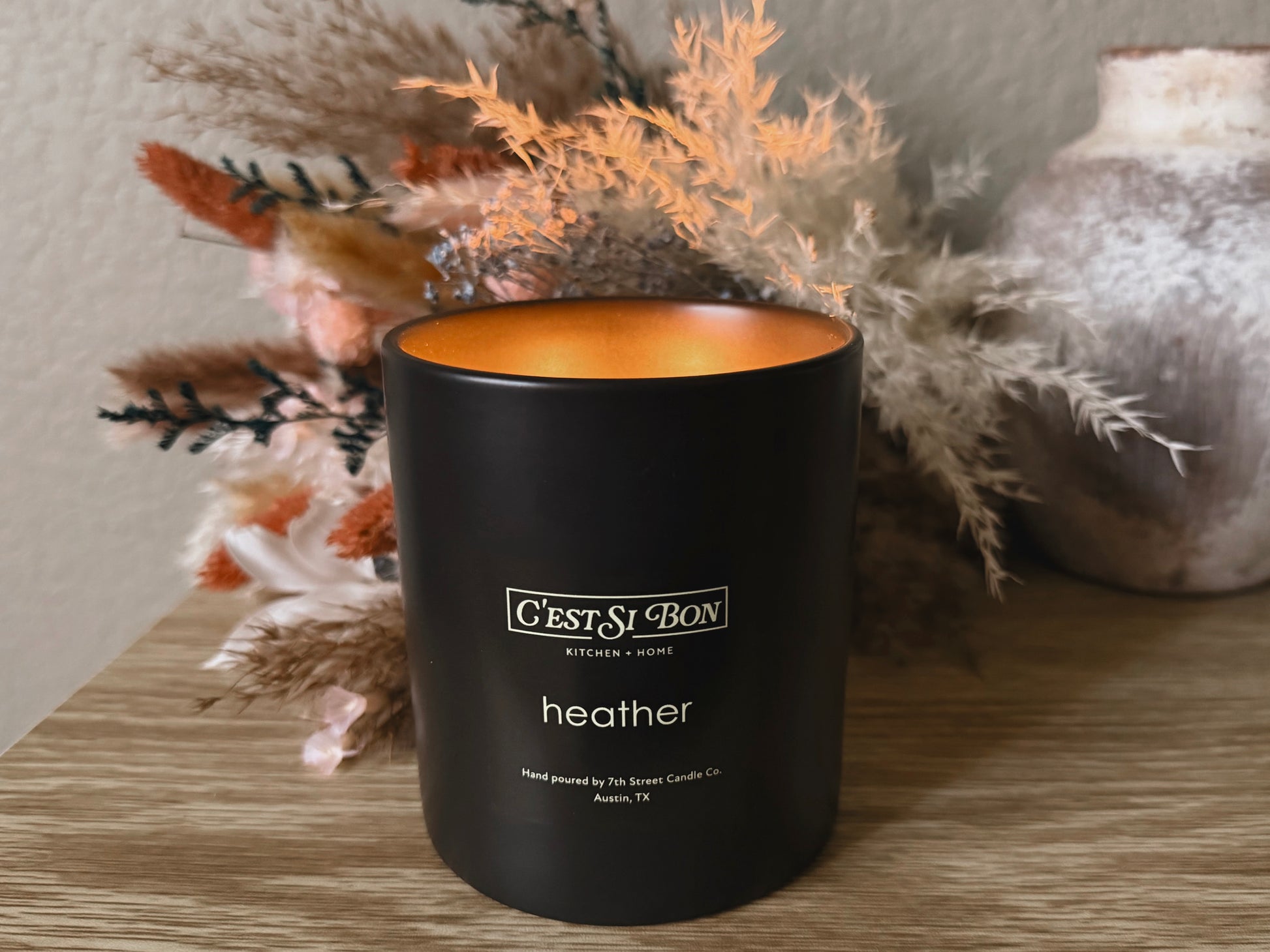 Black candle labeled 'Cést Si Bon' in the scent 'Heather' on a wooden surface with decorative elements.