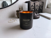 C'est Si Bon-branded black ceramic jar candle in the scent Moi lit on a vanity, with earrings, perfume, a journal, and a mirror in the background. 