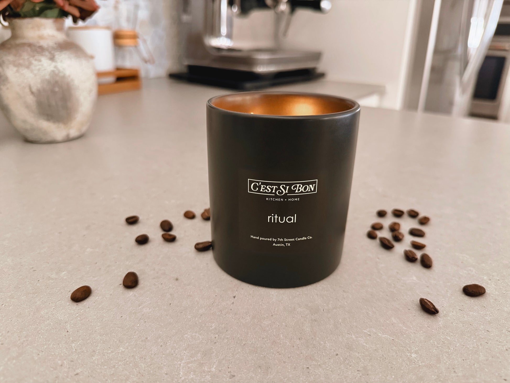 C'est Si Bon-branded black ceramic jar candle in the scent Ritual with coffee beans scattered on a kitchen counter with an espresso machine in the background.