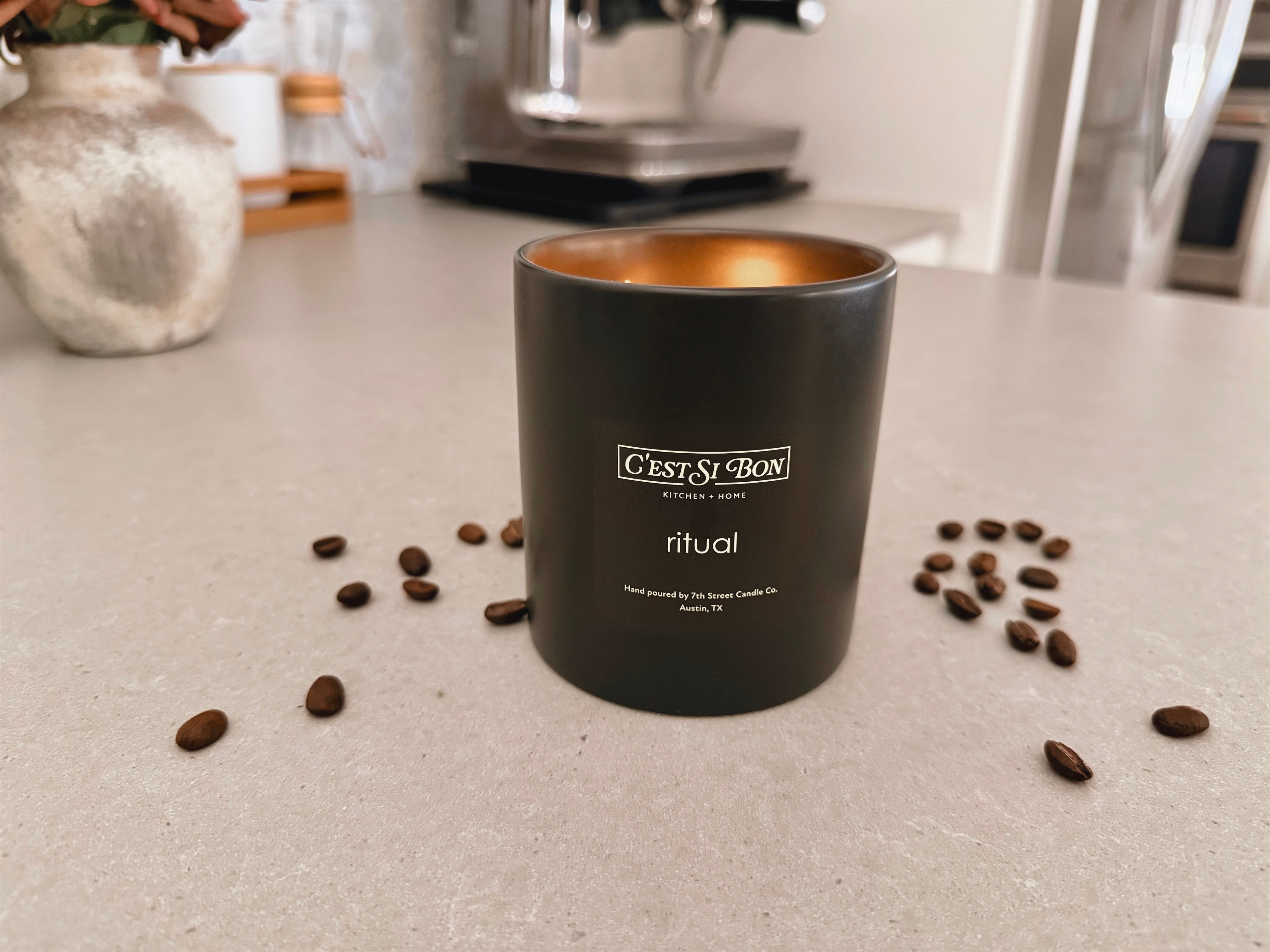 C'est Si Bon-branded black ceramic jar candle in the scent Ritual with coffee beans scattered on a kitchen counter with an espresso machine in the background.