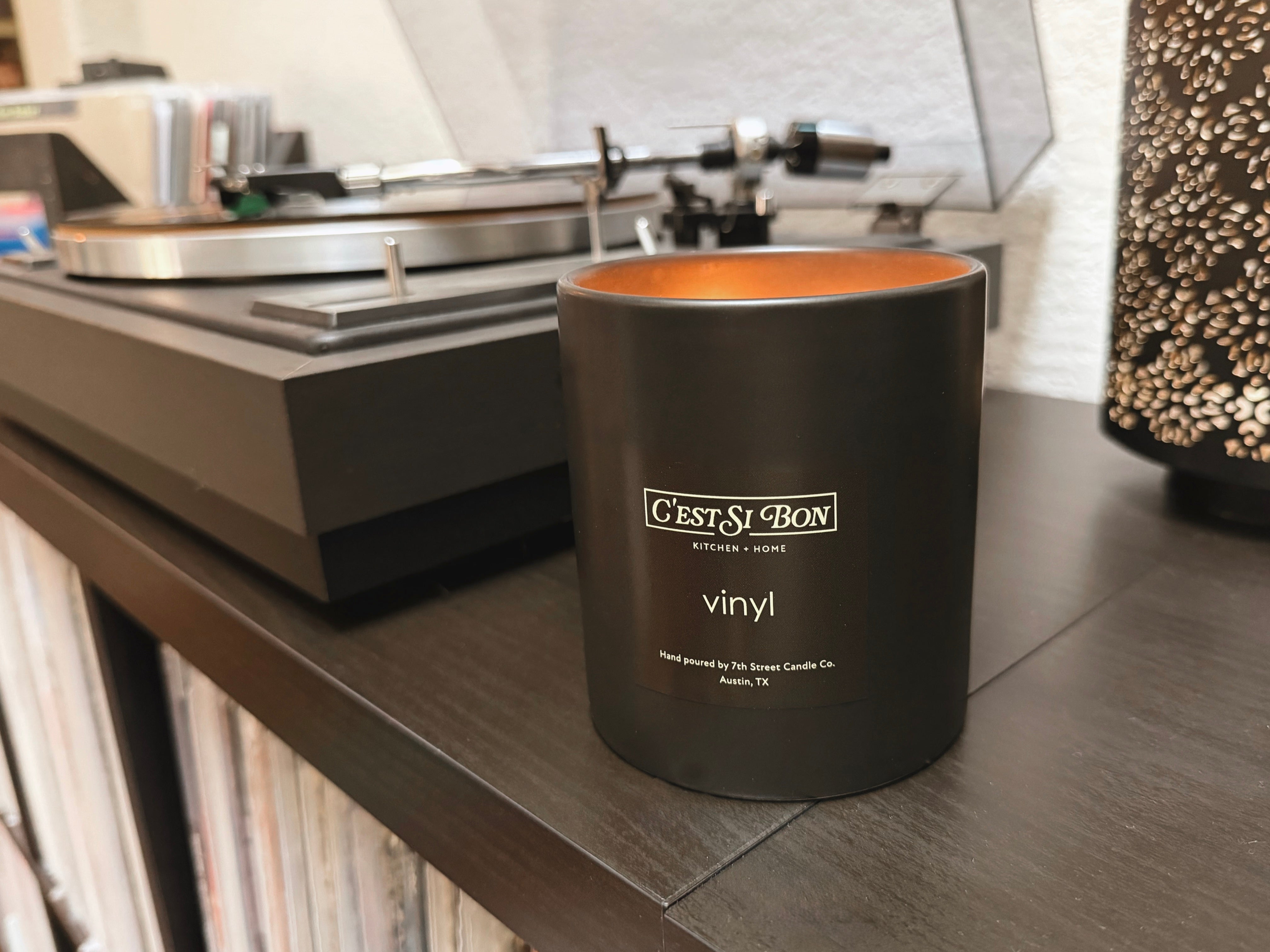 C'est Si Bon branded black ceramic jar candle in the scent Vinyl on a shelf with vinyl records and a record player in the background.